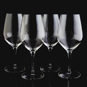 May include: Five clear wine glasses with long stems are arranged in a row against a black background. The glasses have a classic, elegant shape, with a wide bowl and a slender stem. The glasses are empty and reflect the light.