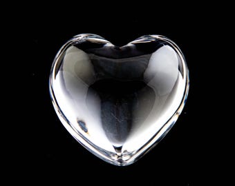 May include: A clear, transparent glass heart-shaped object with smooth, rounded edges and a polished surface. The heart features a concave center that reflects light, creating subtle highlights and shadows across its curved, solid form.