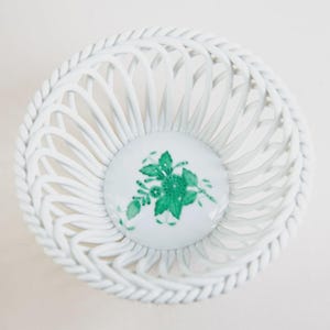 Puede incluir: Cesta de cerámica blanca con diseño tejido y borde trenzado. El centro de la cesta presenta un diseño circular con un motivo floral verde. La cesta probablemente se utiliza con fines decorativos o para guardar objetos pequeños.