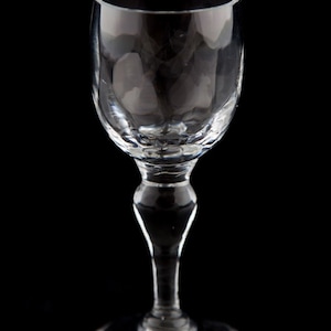 May include: A clear glass shot glass with a stemmed base. The glass has a rounded bowl and a delicate, elegant design.