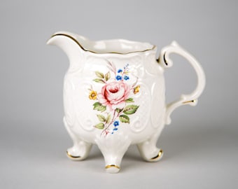 Könnte beinhalten: Ein weißer Porzellan-Milchkännchen mit einem Blumendesign mit einer rosa Rose, blauen und gelben Blumen und grünen Blättern. Der Milchkännchen hat drei goldfarbene Füße.