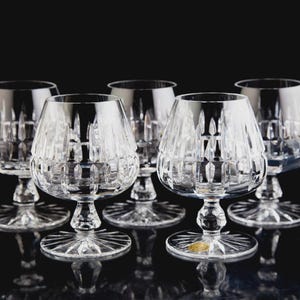 May include: Five clear crystal brandy snifters with a faceted design. The glasses have a rounded bowl, a short stem, and a wide, starburst-patterned base. The background is black, highlighting the clarity and sparkle of the glassware.