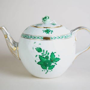 May include: White porcelain teapot with green floral designs and gold trim. The teapot features a rounded body, a curved spout, and a handle. The lid is topped with a green rose-shaped knob. A decorative piece for tea parties or kitchen decor.