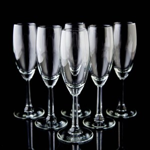 May include: Six clear glass champagne flutes arranged in a row on a black background. The flutes are empty and have a classic design.