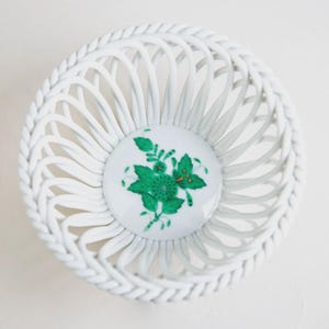 Puede incluir: Cesta de cerámica blanca con un diseño tejido y un borde decorativo en forma de cuerda. El centro presenta un círculo blanco con un diseño floral verde. Esta cesta decorativa es ideal para exhibir objetos pequeños.