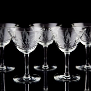 May include: Set of six clear glass cocktail glasses with a delicate etched floral design. The glasses are arranged in a row on a black background.