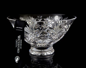 May include: A clear crystal bowl with a cut-glass design. The bowl is on a black background and has a label that reads "House of Waterford Crystal, Made in Ireland".