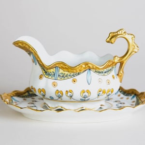 May include: A white porcelain gravy boat and matching saucer with gold trim and floral accents. The gravy boat has a decorative gold handle and a scalloped rim. The design includes yellow, blue, and gold floral patterns.