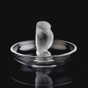 Könnte beinhalten: Eine klare Glas-Eulenfigur steht in einer flachen, kreisförmigen Schale. Die Eule hat geätzte Federdetails. Die Schale und die Eule sind vor einem schwarzen Hintergrund platziert, was einen Kontrast erzeugt.