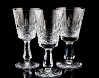 Puede incluir: Tres vasos de chupito de cristal tallado, con tallo, transparentes. Los vasos están dispuestos en fila sobre una superficie negra.