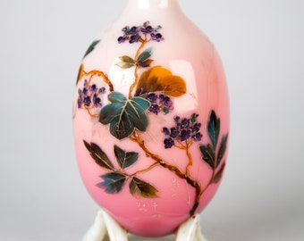 May include: A pink and white glass vase with a floral design. The vase has a rounded body and a small opening at the top. It is sitting on a white stand with three legs.
