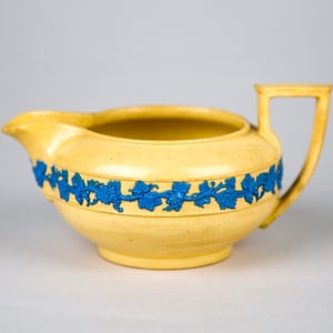 May include: A yellow ceramic creamer with a blue vine design around the top. The creamer has a rounded body and a handle.
