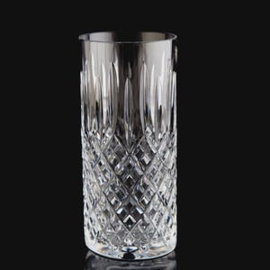 May include: A clear, cut-crystal highball glass. The glass features a diamond pattern on the lower portion and vertical cuts near the rim. The glass is cylindrical and sits on a black surface.