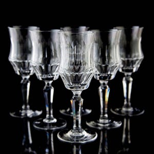 May include: Six clear crystal wine glasses with a tulip-shaped bowl and a long stem are arranged on a black surface. The glasses have a cut design on the bowl and stem. The glasses are reflecting on the black surface.