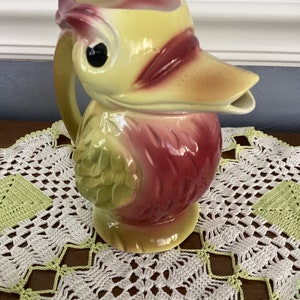 May include: A ceramic pitcher shaped like a bird with a yellow, green, and red glaze. The bird has a large beak and a single black eye.