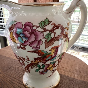 May include: A white ceramic pitcher with a gold rim and a floral design featuring a colorful bird. The pitcher has a wide base and a handle.