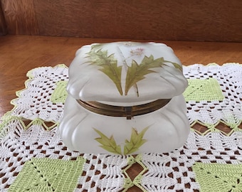 May include: A white frosted glass trinket box with a hinged lid. The box is decorated with a painted design of green leaves and a white flower. The box is sitting on a white and green crocheted doily.