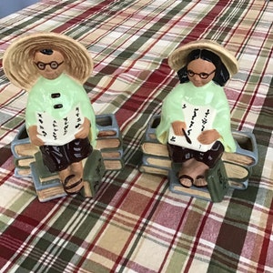 May include: Two ceramic figurines of people wearing conical hats and sitting on stacks of books. The figurines are painted in green, brown, and white. The people are holding books and appear to be reading.
