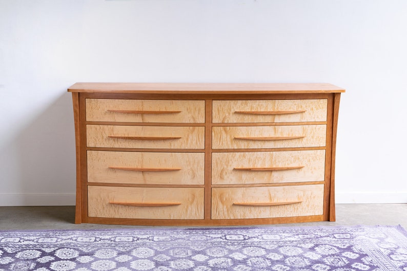 May include: A curved wooden dresser with eight drawers. The dresser is made of light wood with a natural finish. The drawers have simple, curved wooden handles.
