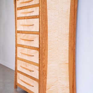 May include: A tall wooden dresser with six drawers and a curved front panel. The dresser is made of cherry wood with a natural finish. The drawers have simple, wooden pulls.