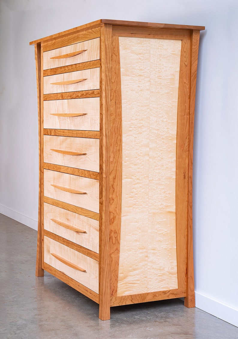 May include: A tall wooden dresser with six drawers and a curved front panel. The dresser is made of cherry wood with a natural finish. The drawers have simple, wooden pulls.
