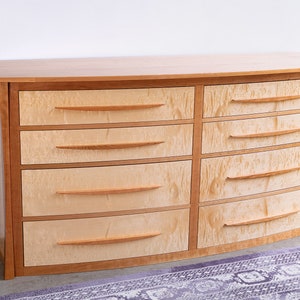 May include: A curved wooden dresser with six drawers. The dresser is made of light wood with a natural finish. The drawers have simple, curved wooden handles.