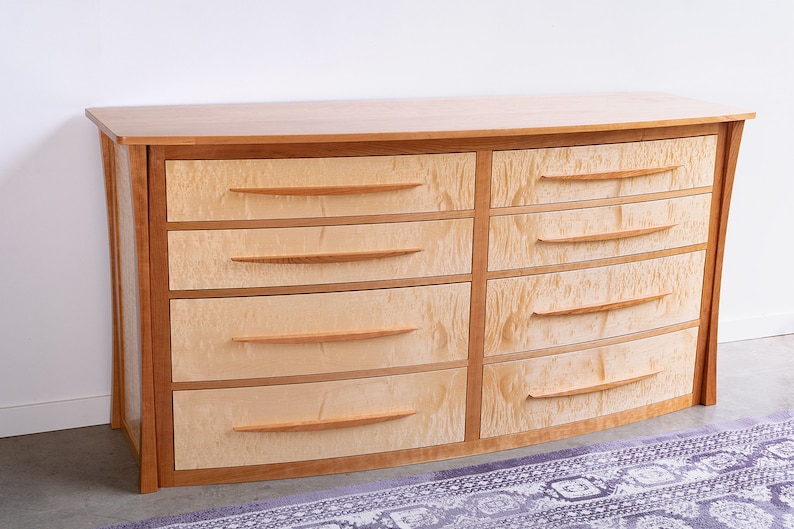 May include: A curved wooden dresser with six drawers. The dresser is made of light wood with a natural finish. The drawers have simple, curved wooden handles.