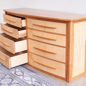 May include: A curved wooden dresser with six drawers, all of which are open. The dresser is made of light wood with a natural finish. The drawers have a light wood interior and are made with dovetail joints. The dresser has a decorative inlay on the side.