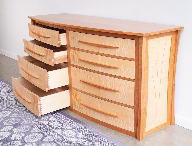 May include: A curved wooden dresser with six drawers, all of which are open. The dresser is made of light wood with a natural finish. The drawers have a light wood interior and are made with dovetail joints. The dresser has a decorative inlay on the side.