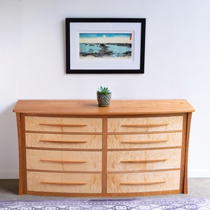 May include: A curved wooden dresser with six drawers. The dresser is made of light wood with a natural finish. The drawers have a light wood finish with a darker wood trim. A small potted plant sits on top of the dresser.