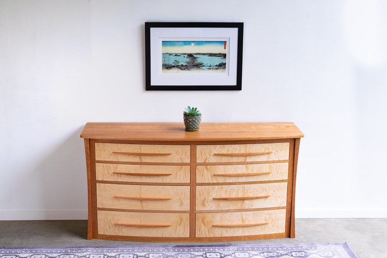 May include: A curved wooden dresser with six drawers. The dresser is made of light wood with a natural finish. The drawers have a light wood finish with a darker wood trim. A small potted plant sits on top of the dresser.