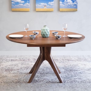 May include: A round wooden dining table with a unique, sculptural base. The table is set with two place settings, each with a white plate, a fork, and a wine glass. There is a vase with a light green textured finish in the center of the table.