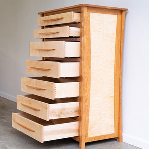 May include: A tall wooden dresser with seven drawers, all open and revealing the light wood interior. The dresser is made of cherry wood with a natural finish and has a curved front.