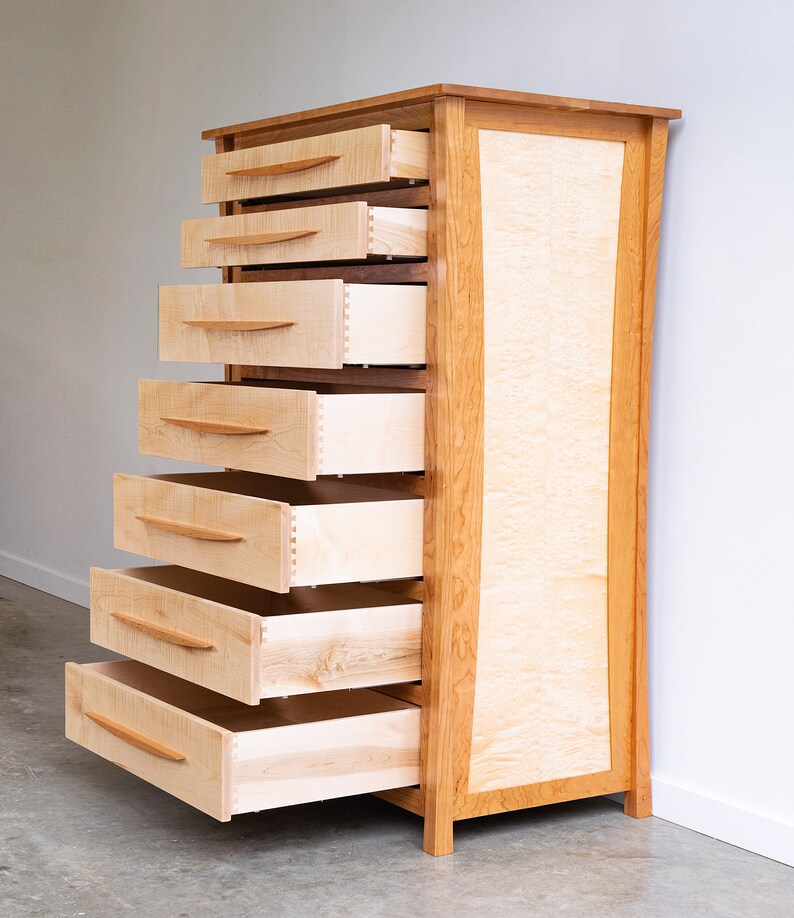 May include: A tall wooden dresser with seven drawers, all open and revealing the light wood interior. The dresser is made of cherry wood with a natural finish and has a curved front.