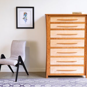May include: A light wood dresser with six drawers and a framed print on the wall behind it. A chair with a light gray fabric seat and black legs is in the foreground.