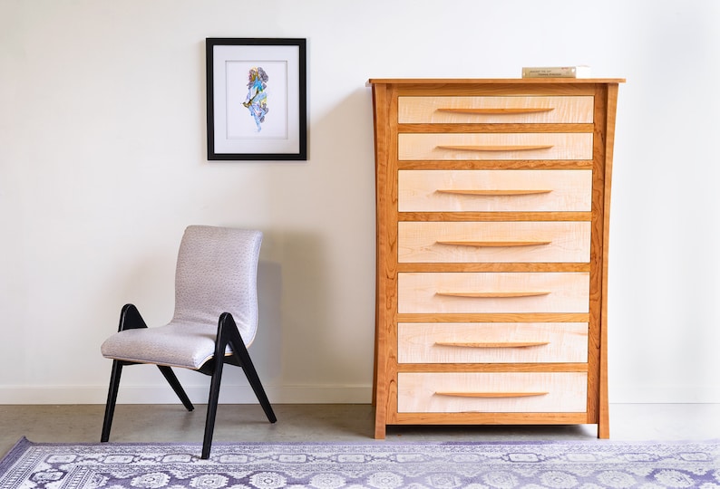 May include: A light wood dresser with six drawers and a framed print on the wall behind it. A chair with a light gray fabric seat and black legs is in the foreground.