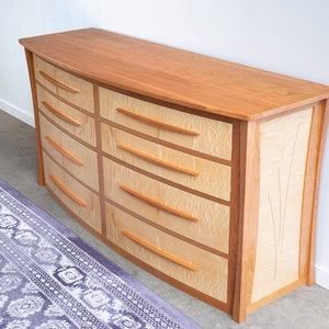 May include: A curved, wooden dresser with six drawers. The dresser is made of cherry wood and has a light wood finish. The drawers have a light wood finish and are accented with a darker wood trim. The dresser has a curved front and a flat top.
