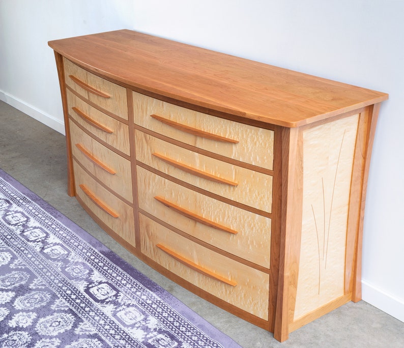 May include: A curved, wooden dresser with six drawers. The dresser is made of cherry wood and has a light wood finish. The drawers have a light wood finish and are accented with a darker wood trim. The dresser has a curved front and a flat top.