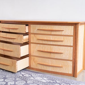 May include: A light wood dresser with six drawers, three of which are open. The drawers have curved fronts and wooden handles. The dresser has a curved front and is made of wood with a natural finish.