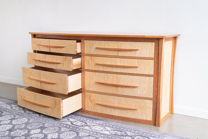 May include: A light wood dresser with six drawers, three of which are open. The drawers have curved fronts and wooden handles. The dresser has a curved front and is made of wood with a natural finish.