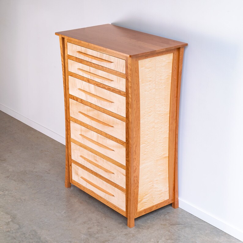 May include: A six-drawer dresser made of light wood with a natural wood finish. The drawers have a simple design with a slight curve at the top. The dresser has a curved front and sides.