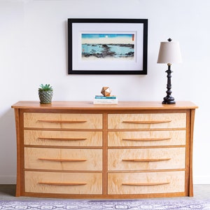 May include: A light wood dresser with six drawers and curved edges. The dresser has a natural wood finish and is decorated with a small potted plant, a geometric object, and a framed print.