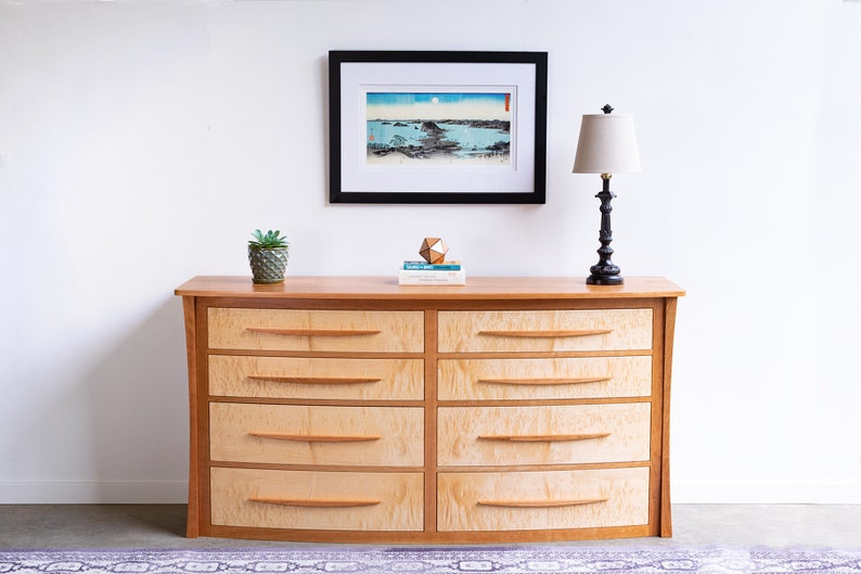May include: A light wood dresser with six drawers and curved edges. The dresser has a natural wood finish and is decorated with a small potted plant, a geometric object, and a framed print.
