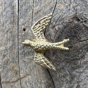 May include: A gold metal bird figurine in flight, with wings spread wide, mounted on a weathered wooden surface.