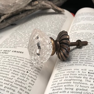 Pode incluir: Um puxador de móvel em vidro transparente facetado com formato de diamante e uma base de metal em tom de bronze antigo escuro com design canelado. O puxador inclui um parafuso de metal roscado e uma porca correspondente para instalação.