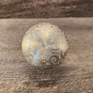 May include: A white sphere with a gold swirl pattern. The sphere is on a wooden surface.