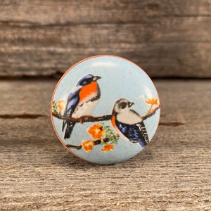 May include: A ceramic knob with a blue background and two blue birds with orange accents perched on a branch with orange flowers.