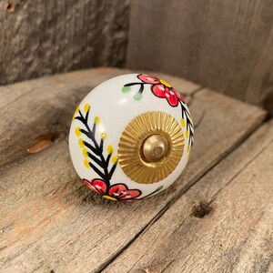 May include: White ceramic knob with a floral design featuring red, yellow, and green flowers. The knob has a gold metal center.