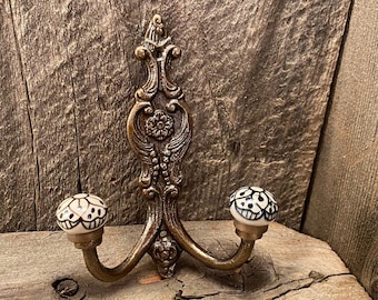 May include: Ornate brass wall hook with two curved arms and decorative floral and scrollwork details. The hook features two white ceramic knobs with black floral patterns. A vintage-style decorative piece.