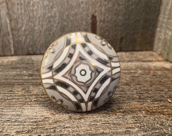 Może przedstawiać: Okrągły ceramiczny gałka z geometrycznym wzorem w kolorze czarnym, białym i złotym. Gałka ma wypukły wzór i gładkie wykończenie.